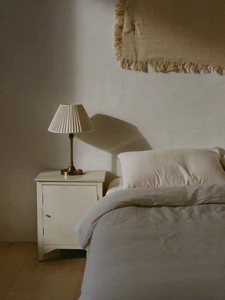 A bed in flax linen beside an ivory nightstand with a brass lamp