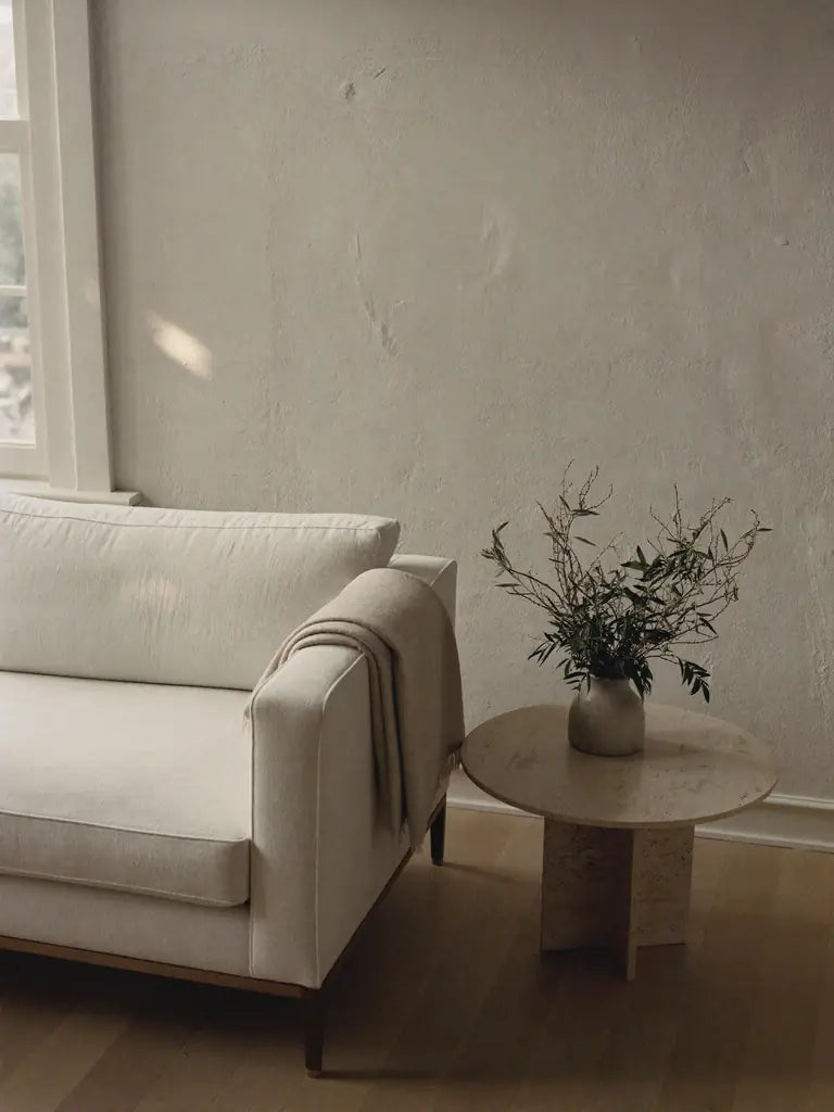 The arm of a linen sofa beside a round travertine side table